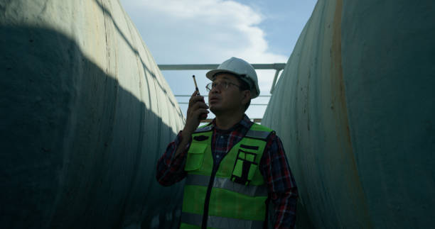 istock Senior maintenance engineer wearing a reflective vest and using a walkie-talkie while working with a blueprint drawing at a large water tank in a construction site of the factory. 2168109728