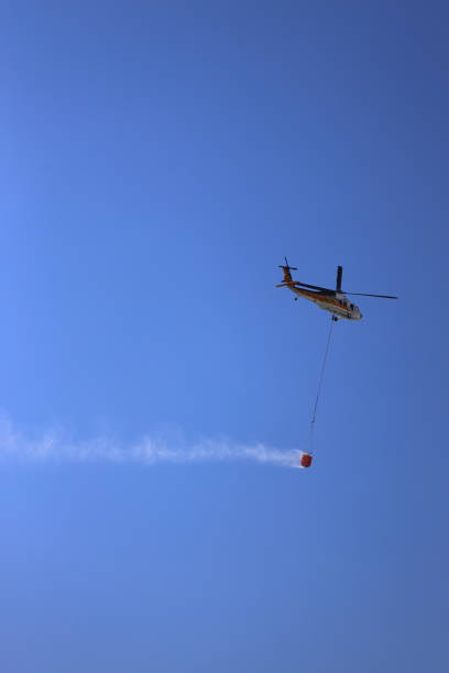 Badajoz, Spain. June 17, 2025. A yellow Eurocopter firefighting helicopter flies through smoke, deploying its water bucket to extinguish a raging wildfire.