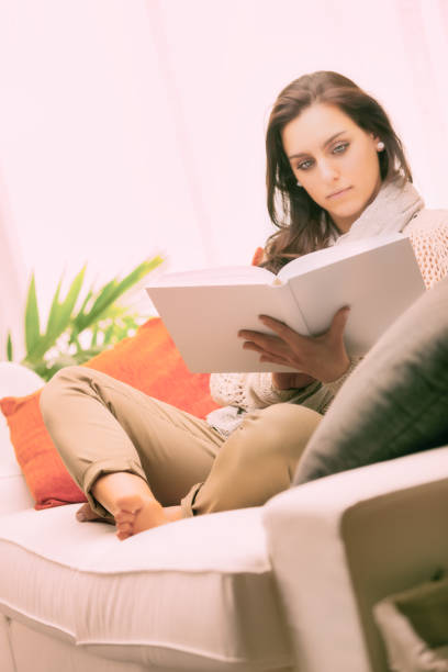 istock Beautiful young woman reading a book on her couch 2168141085