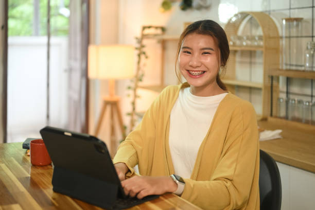 istock Smiling young asian lady in yellow cardigan sitting at a kitchen counter and using digital tablet 2168155771
