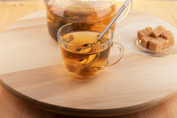 Large leaf tea is brewed in a glass teapot. Hot tea is in a glass cup nearby, and brown lump sugar is on a glass saucer on a round wooden tray