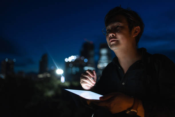 istock Man with glasses focused on his tablet while standing on a rooftop at night, with city skyscrapers lit up in the background. 2168160459