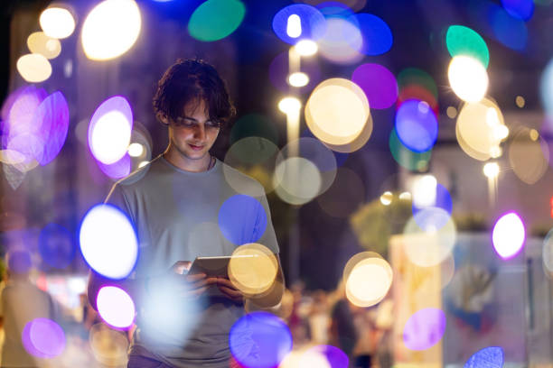 Teenage boy standing and holding tablet, looking at device screen, through the city night lights