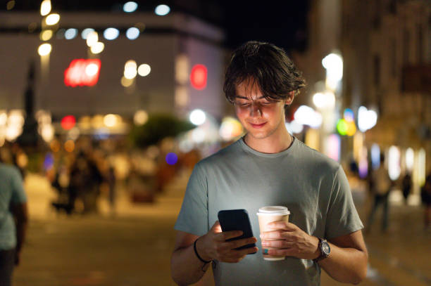 Teenage boy drinking coffee in a cup and using phone in the city at night