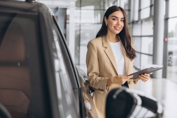 istock Handsome car dealership female worker in suit is holding a folder and smiling while standing near the car 2168173363