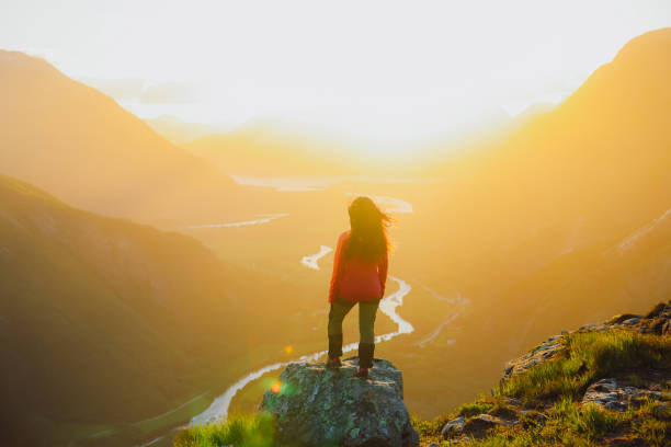 Rear view of active confident sport woman explorer got to the top of the mountain overlooking scenic mountain valley with river from above during very bright colourful summer sunset in Romsdal, Western Norway, Scandinavia