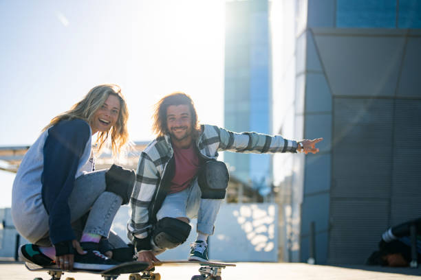 Smiling young man and woman ride skateboards in an urban setting, expressing joy and togetherness, with casual attire suitable for their sporty activity.