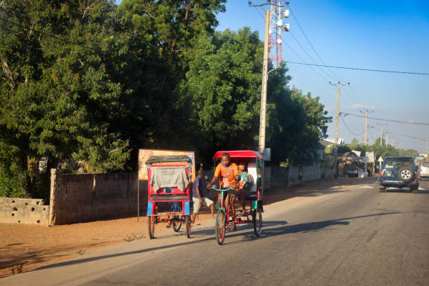 Morondava, Madagascar - May 27, 2024: In Morondava, Madagascar, rickshaw drivers transport passengers along a busy road, with shops and trees lining the route in the warm afternoon sunlight.