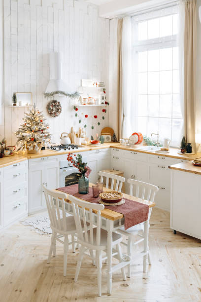 istock Festive dining table in a cozy kitchen featuring a pie and centerpiece, surrounded by Christmas decorations, a small frosted tree with red ornaments, white cabinets, and wooden countertops 2168223634