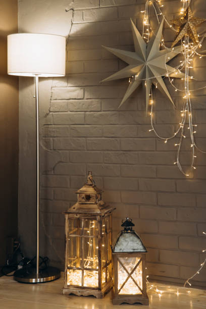 Rustic holiday decor with two lanterns filled with string lights, standing next to a modern floor lamp and star ornaments against a brick wall for a cozy Christmas ambiance