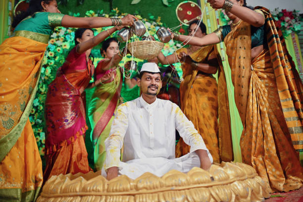 Groom surrounded by family women performing rituals during Haldi Ceremony This image captures an Indian Haldi ceremony, where the groom is surrounded by women from his family who are performing thread rituals. The groom is seen in traditional white kurta sit at the center of the gathering, while his female relatives, dressed in traditional yellow sari lovingly perform sacred rituals. haldi ceremony stock pictures, royalty-free photos & images