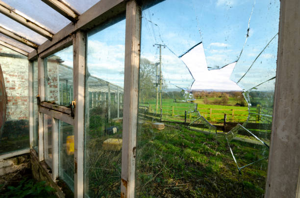 The image shows a broken window in an abandoned greenhouse on a farm in England, offering a view of the surrounding rural landscape.