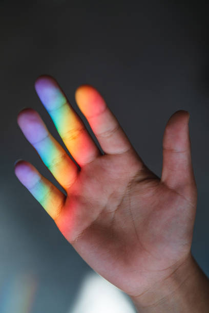 Close-up of a human hand with rainbow colors projected onto the fingers, creating a colorful spectrum effect on the skin.