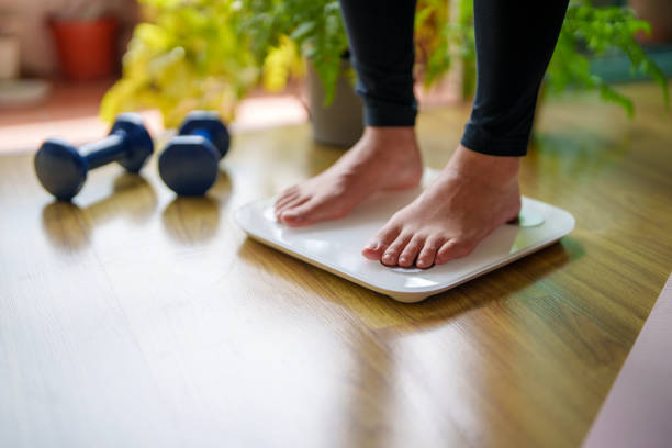 A close-up view of a young woman standing on a weighing scale A close-up view of a young woman standing on a weighing scale, focusing on her feet and the display as she checks her weight. The scene highlights the importance of health and self-awareness in maintaining a balanced lifestyle. low quality of life stock pictures, royalty-free photos & images