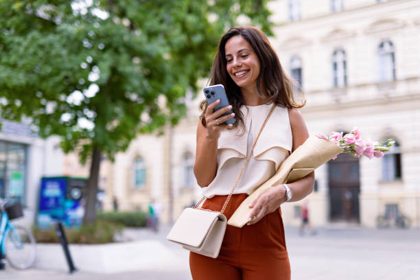istock Woman smiling outdoors holding flowers and using phone 2168360617