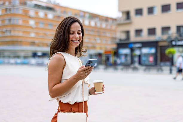 istock Smiling woman checking phone while holding coffee outside. 2168361125