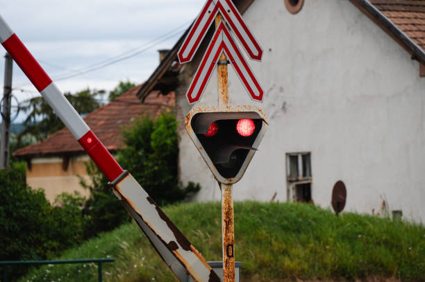 A close-up of a railway crossing signal with red lights illuminated and a partially lowered barrier. The signal post shows signs of rust, adding a rustic touch to the scene, set against a backdrop of an old building and greenery.