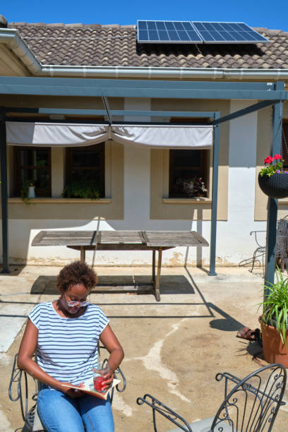 A woman is sitting in a sunny backyard, peacefully reading a book while holding a drink. The backyard features solar panels on the roof and potted plants, creating a serene and eco-friendly atmosphere.
