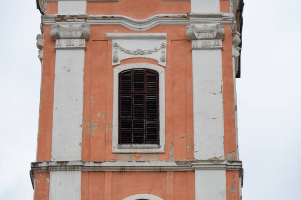A weathered facade of a historical building featuring closed wooden shutters on an arched window. The peeling paint and ornate architectural details highlight the age and character of the structure, evoking a sense of faded grandeur.
