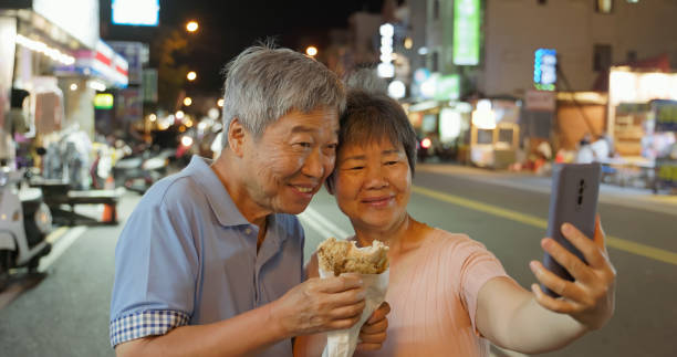 Closeup side view asian elderly couple selfie and video call while eating fried chicken at night market in Kenting Street Taiwan