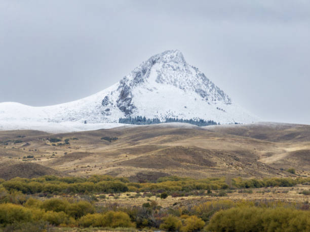 istock Snow-covered peak rising from an arid landscape with sparse vegetation, under a cloudy sky. 2168385293