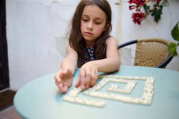 istock Young girl with focused expression playing dominoes on a patio table, engaging in a thoughtful and strategic game in an outdoor setting with a relaxed atmosphere. 2168393689