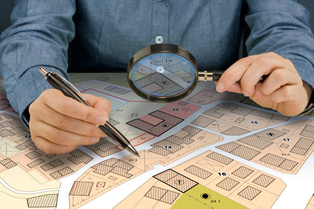 istock SEARCHING BUILDING PLOT FOR HOME CONSTRUCTION - Hand with magnifier on fictional cadastral map and General Urban Plan 2168402678