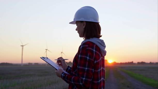 istock Woman engineer is taking notes on a clipboard on a field with wind turbines, as the sun sets in evening. Clean energy and engineering 2168427326