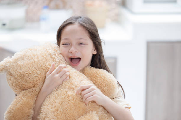 A young girl of African decent, sits up on an exam table at the doctors as she grips her teddy bear tightly.  She is dressed casually and smiling.