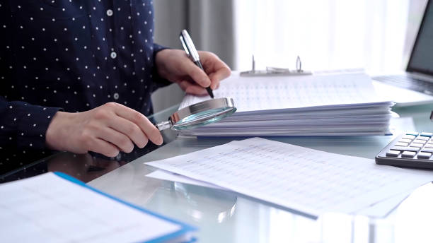istock Businesswoman using calculator and reviewing ring folder of financial documents with magnifying glass at desk in modern office. Audit and taxes in business 2168455251
