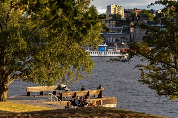 Stockholm, Sweden Aug 16, 2024 People sitting next to the water on Skeppsholmen island, the Baltic Sea, and a city ferry.