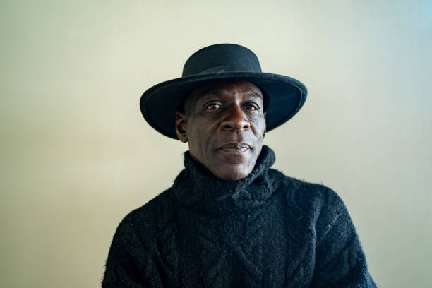 A senior Afro-Caribbean man is looking at the camera, wearing a black cowboy hat and a black sweater against a gradient grey to white background
