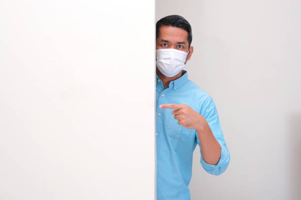 A man wearing medical face mask peeking behind white board and pointing on it