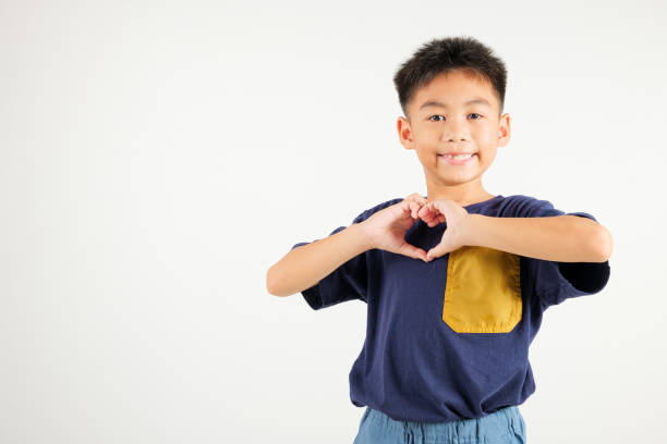 A smiling Asian young kid boy shows confidence, creating a heart shape with fingers and hands on white background. Portrait of primary child sending love for Valentine's A smiling Asian young kid boy shows confidence, creating a heart shape with fingers and hands on white background. Portrait of primary child sending love for Valentine's Day cute boy show love symbol hand stock pictures, royalty-free photos & images