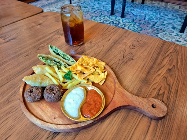 Yummy Snack Mix Platter With Fried Sambosa Cheese Samosa, Mutabbaq, Falafel, Tortilla Chips And French Fries. Serving On A Wooden Tray With Spicy Sauce And Mayonnaise Sauce. Fresh Tea Iced In Glass. Food And Drink Menu. delicioso plato de mezcla de bocadillos con queso sambosa frito, samossa, mutabbaq, falafel, chips de tortilla y papas fritas. sirviendo en una bandeja de madera con salsa picante y mayonesa. té fresco helado en vaso. menú de comida y bebida.