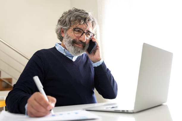 istock Happy mature man working with laptop talking on the phone at home office taking notes. 2168518453