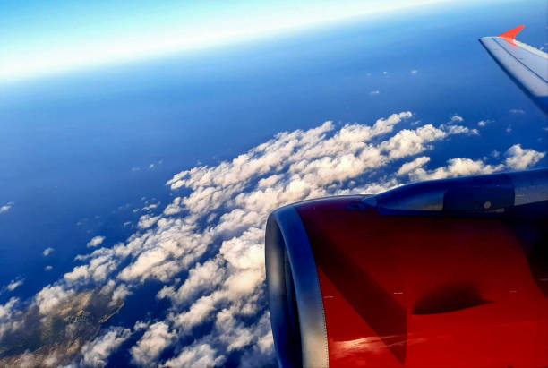 Panoramic view from an airplane window on a sunny day, showcasing a vast expanse of clear blue sky, scattered fluffy clouds, and a landscape stretching to the horizon. The sunlight illuminates the scene, highlighting the intricate patterns of the land below and the serene beauty of the aerial perspective.
