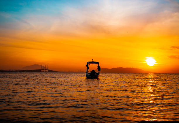 In the warm embrace of the sunset, the Penang Bridge stands tall, connecting the island to the mainland, a route taken by countless travelers daily. Yet, in the quiet waters below, a lone boat drifts, silhouetted against a breathtaking sunset, symbolizing a peaceful escape from the hustle and bustle