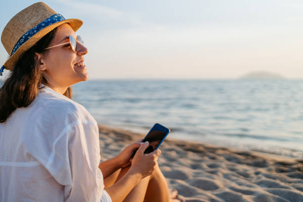 istock Young Woman Using Her Phone While Sitting On The Beach In Neos Marmaras At Sunset 2168568893