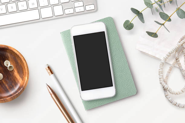 bright feminine smart phone mock up with mint notebook, eucalyptus twigs, accessories and a wooden bowl on a white boho styled desk. Flat lay, top view