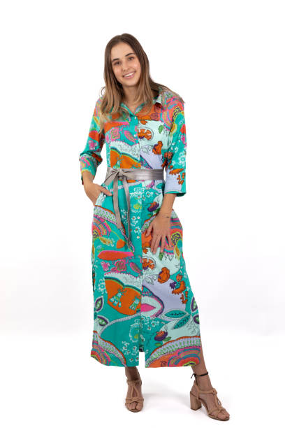 A young woman stands in a full-length image, wearing a brightly colored and bold patterned shirt dress against a crisp white background