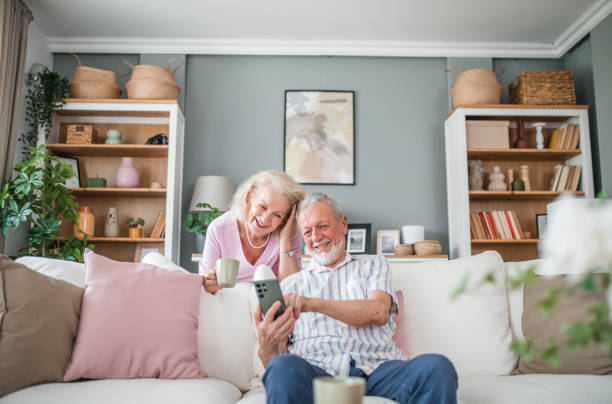 istock A happy senior man is sitting on a sofa and having fun using a mobile phone, while his wife is standing behind him holding a cup of coffee in her hands 2168604527
