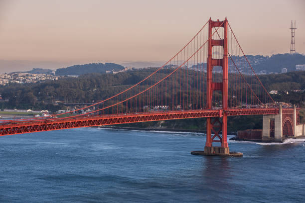 As the famous Golden Gate Bridge crosses the Golden Gate strait, we can see a calm sunset warming the house-filled hills in the background.