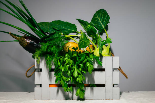 Assorted fresh vegetables in a rustic wooden crate against a grey backdrop.
