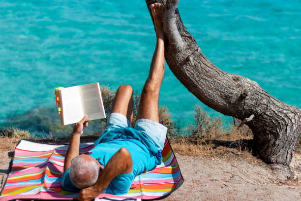 An elderly man is reading a book while sitting in a folding chair on a pebble beach on a hot day