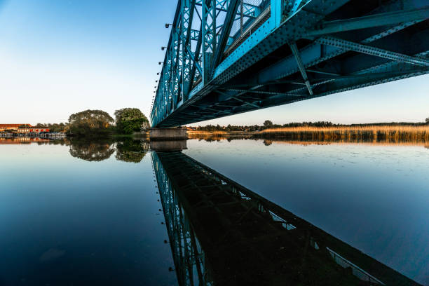 Underneath the blue bridge towering over a river near wildlife marsh at between blue hour and golden hour with still water reflection of bridge POV