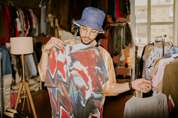 Person examining vibrant tie-dye shirt in cozy clothing store with a large window in background showcasing natural light. Shelves of various garments and unique decor surround them