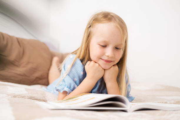 Adorable little girl wearing eyeglasses reading a book in white living room on beautiful summer day