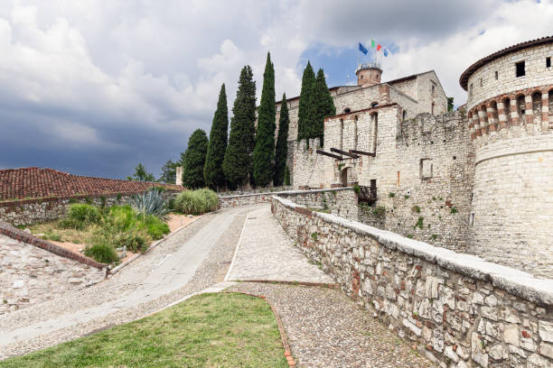 Brescia - Italy. June 26, 2024: Brescia Castle in Italy features flags on the main tower, a medieval drawbridge, stone fortifications, and tall cypress trees lining a cobblestone pathway under a moody cloudy sky