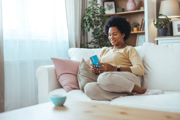 istock A smiling elegant African-American female using her smartphone while sitting on the cozy sofa in the living room. 2168743407